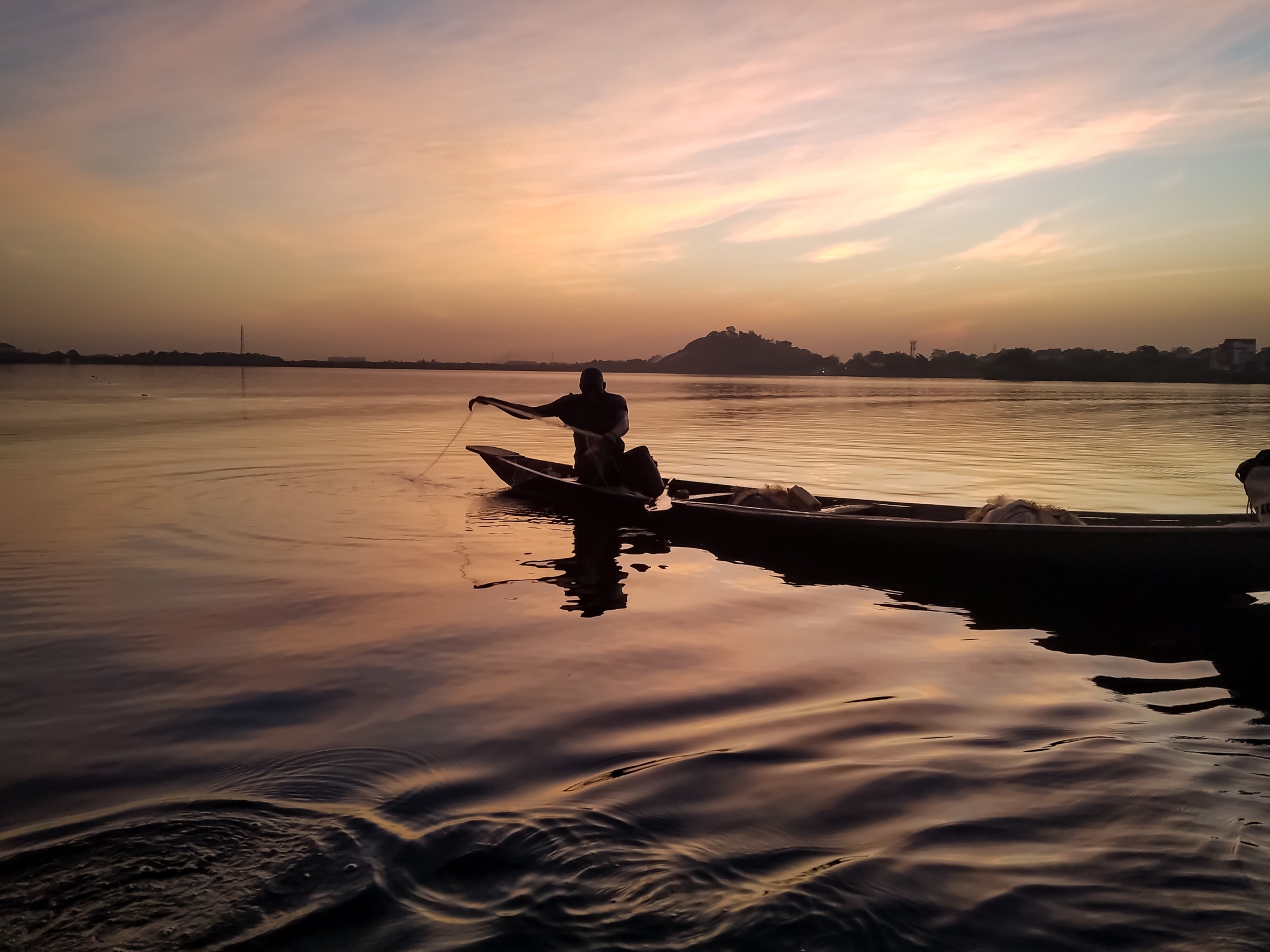 A fisherman casting a net from a wooden canoe on a Nigerian river at dusk